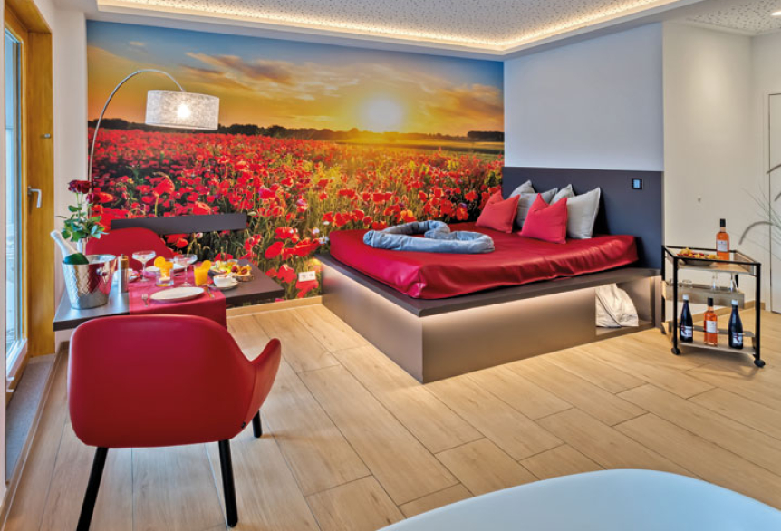 Ausschnitt aus dem Privat Spa. Ein Wasserbett mit roten Dekokissen und einer grauen Kuscheldecke. Das Bett wird von unten mit einem Lichtschlauch warm gelb beleuchtet. Auf der Hintergrundwand ist ein Sonnenuntergang zu sehen und ein Kornblumenfeld in Rot abgebildet. Zwei rote Sesselartige Stühle mit einem gedeckten Tisch.
