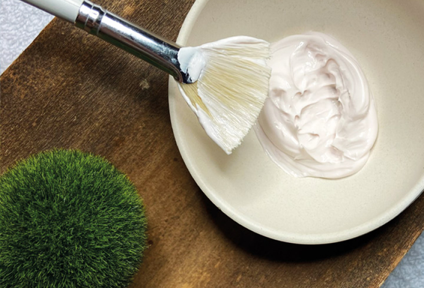 Schale mit cremiger Gesichtsmaske und Pinsel zum Auftragen, dekorativ auf einem Holztablett arrangiert, neben einer kleinen grünen Mooskugel.