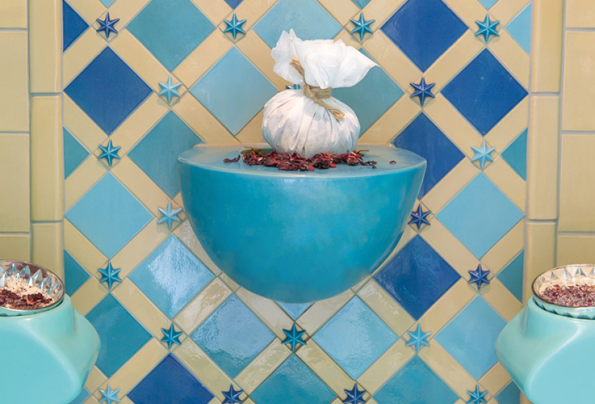 Blick auf einen stilvoll gestalteten orientalischen Rhasul mit türkisfarbenen Sitzflächen und einer kunstvoll gefliesten Wand in Blau- und Beigetönen. In der Mitte befindet sich ein dekorativer Dampfbrunnen.