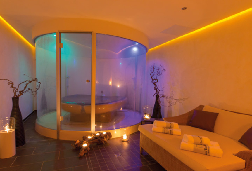Stimmungsvolles Spa-Zimmer mit runder Glaskabine für Anwendungen, dekorativen Vasen, Kerzenlicht und einer gemütlichen Liege mit aufgerollten Handtüchern.