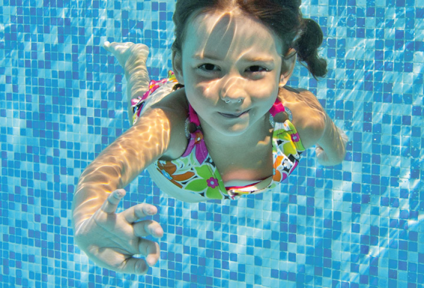 Unterwasseraufnahme eines kleinen Mädchens in einem bunten Badeanzug, dass das Schwimmen lernt. Sie lächelt und winkt in Richtung Kamera.