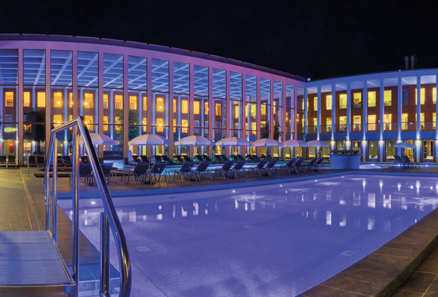 Beleuchteter Außenbereich der SaarowTherme bei Nacht: Modernes Gebäude mit großer Fensterfront, farbiger Lichtakzenten, leerem Solebecken im Vordergrund und aufgereihten Liegestühlen mit Sonnenschirmen.