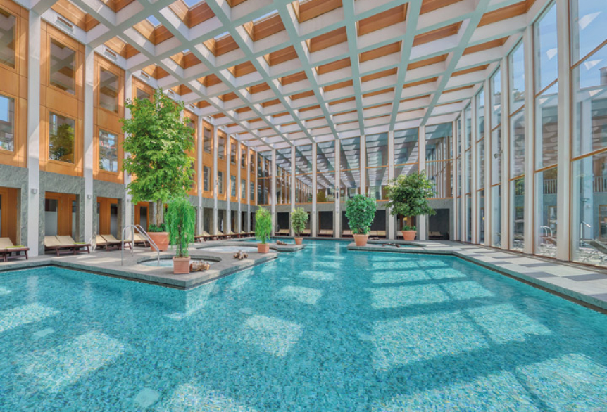 Lichtdurchflutetes Thermalbad mit großen Fenstern und Deckenkuppel. Türkisblaues Wasser und grüne Pflanzen vermitteln ein Ort zum Entspannen und Erholen.