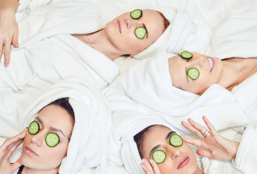 Vier Frauen in weißen Bademänteln und Handtuchturbanen liegen entspannt nebeneinander, mit Gurkenscheiben auf den Augen. Ein klassischer Spa-Moment zur Entspannung und Hautpflege.