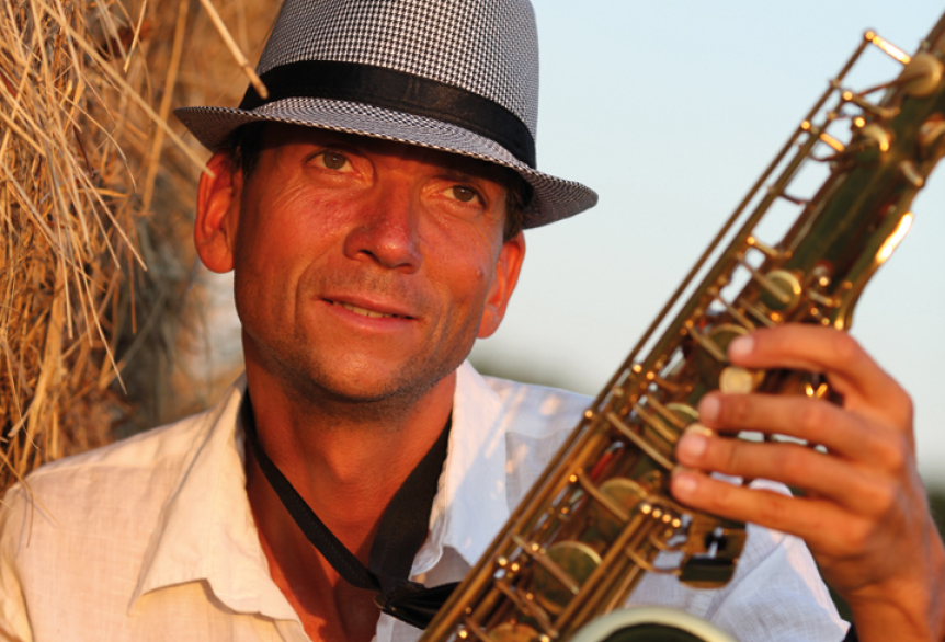 Mann mit hellem Hemd und grauem Hut sitzt entspannt im warmen Abendlicht vor einer Strohwand und hält ein Saxophon in der Hand – eine stimmungsvolle Szene mit musikalischem Flair.