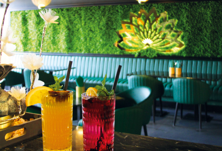 Zwei farbenfrohe Cocktails – einer gelb, einer rot – mit Minze und Zitronenscheiben dekoriert, stehen auf einem Tisch in einer stilvoll eingerichteten Lounge mit grün gepolsterten Sitzmöbeln und einer leuchtenden Pflanzenwand im Hintergrund.