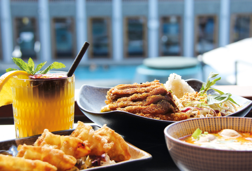 Drei Teller mit asiatisch inspirierten Speisen und ein gelber Cocktail mit Minze stehen auf einem Tisch mit Blick auf ein Thermenaußenbecken. Im Hintergrund entspannen Gäste im Wasser, umgeben von moderner Architektur und Sonnenlicht.