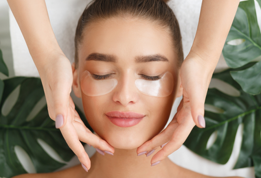 Eine Junge Frau erhält eine Beautybehandlung. Auf dem Rücken liegt Sie auf eine Wellnessliege. Ihr Gesicht mit Augenpads bestückt. Ihre Haare in einem Bun hochgesteckt. Die Hände der Therapeutin streicheln sanft Ihr Gesicht mit pflegenden Produkten. 