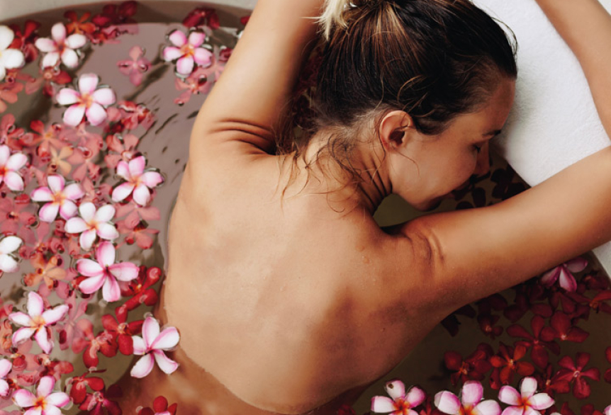 Eine Frau entspannt in einer runden Badewanne mit warmem Wasser und schwimmenden Blüten. Sie lehnt sich mit geschlossenen Augen und erhobenen Armen an den Wannenrand, umgeben von Wellnessprodukten. Die Atmosphäre wirkt ruhig und erholsam.