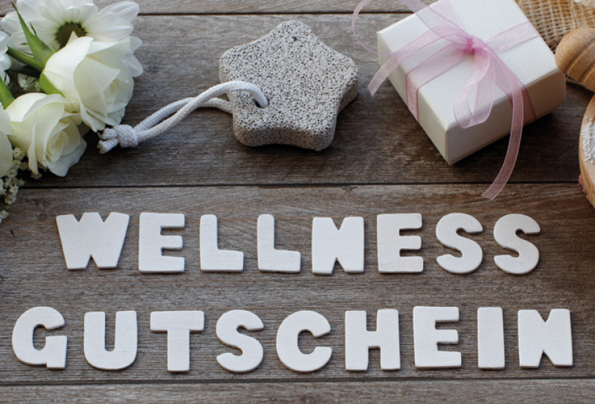 Imagebild mit dem Schriftzug ‚Wellness Gutschein‘ in Weiß im Vordergrund. Im Hintergrund Dekoelemente: weiße Rosen, ein sternförmiger Bimsstein und eine kleine Geschenkbox mit rosa Stoffband. 