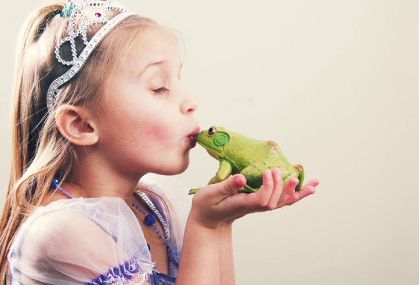 Ein Kind in einem Prinzessinnenkostüm mit Krone hält einen grünen Frosch in den Händen und küsst ihn. Die Szene erinnert an das Märchen „Der Froschkönig“ und wirkt verspielt und fantasievoll.
