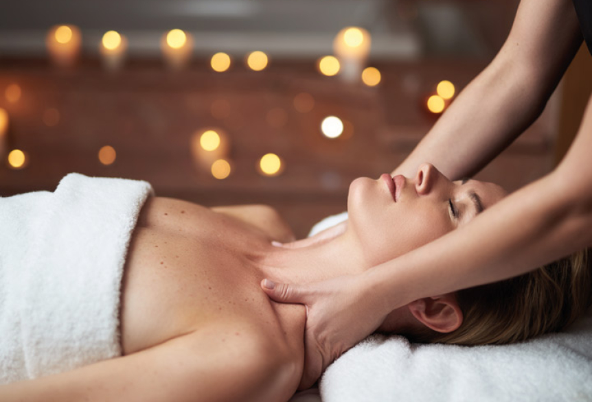 Frau liegt auf dem Rücken auf eine Massageliege eingewickelt in einem Handtuch. Am Kopfende der Massageliege steht eine Masseurin und massiert die Schulter der Frau. 