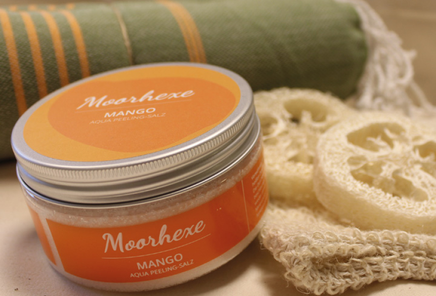 Nahaufnahme eines Peelingsalzes der Marke Moorhexe mit orangem Etikett und der Aufschrift ‚Moorhexe Mango. Im Hintergrund Seifenschwämme und ein grünes Hamamtuch.