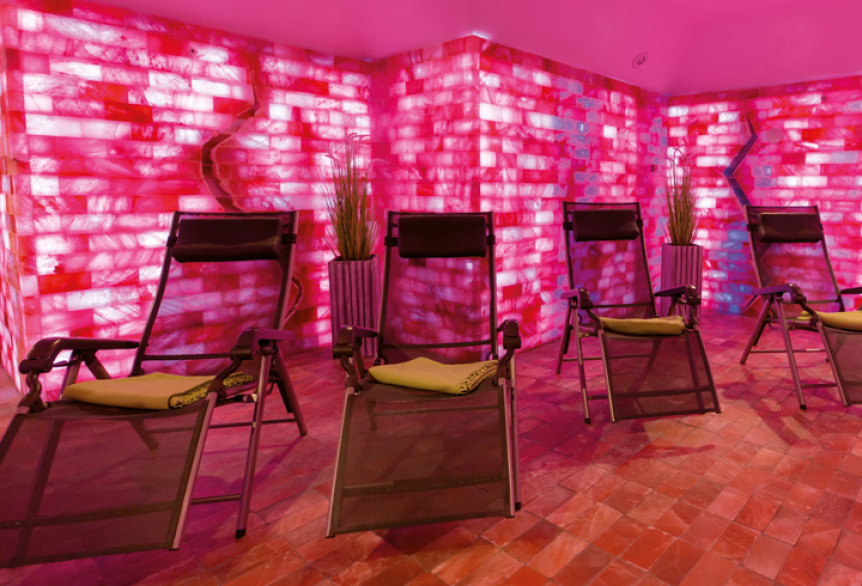 Mehrere Relaxliegen in einem Ruheraum mit roten Salzwänden, warmem rosa Licht und salzbedecktem Boden; dekorative Pflanzen an den Wänden.