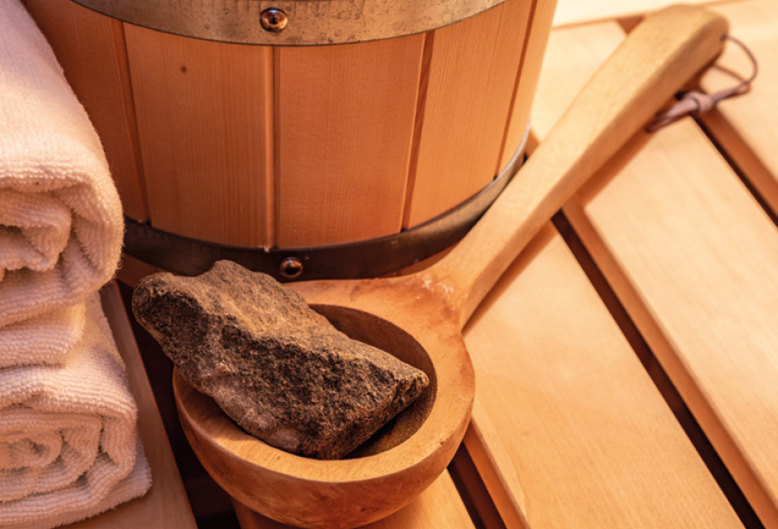 Saunakelle mit einem Saunastein liegt neben einem Saunaeimer und zusammengerollte Handtücher. 