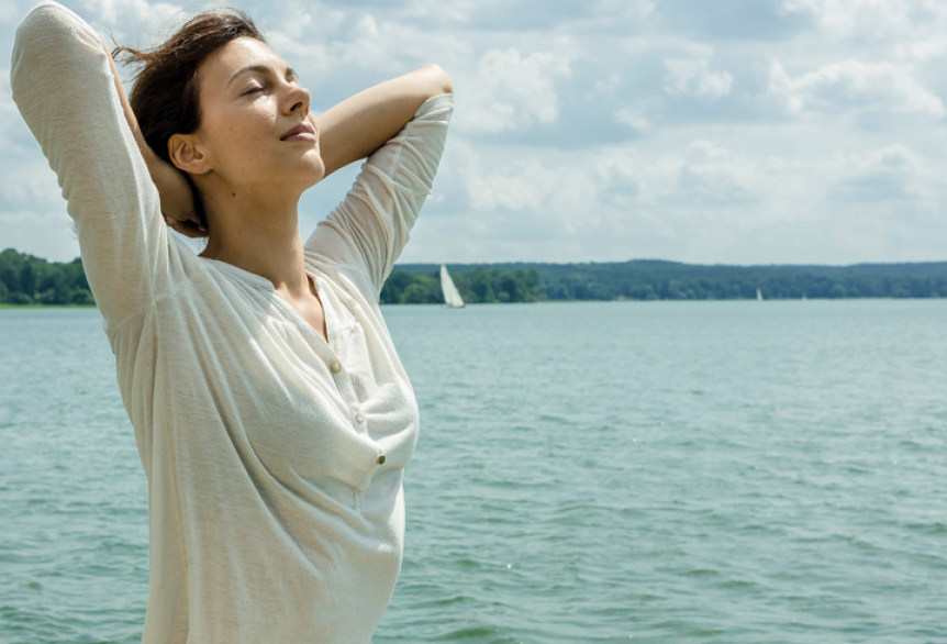 Eine Person steht entspannt am Wasser, hebt die Arme hinter den Kopf und schließt die Augen. Im Hintergrund sind ein See, Bäume und Segelboote unter einem leicht bewölkten Himmel zu sehen – die Szene vermittelt Ruhe und Wohlbefinden.