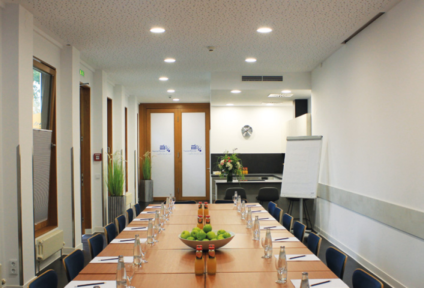 Tagungsraum mit langem Tisch, blauen Stühlen und großem Bildschirm mit dem Logo der SaarowTherme. Auf dem Tisch stehen Wasserflaschen, Orangensaft, Schreibblöcke und Bleistifte, vorbereitet für eine Tagung. Im Hintergrund ist die integrierte Küche des Tagungsraums zu sehen.