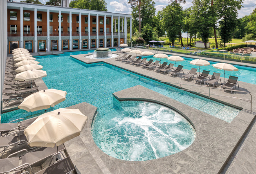 Außenbereich der SaarowTherme in Bad Saarow mit Thermalsolebecken, großem Whirlpool sowie Liegestühlen und weißen Sonnenschirmen am Beckenrand. Im Hintergrund liegt der grüne Kurpark direkt am Scharmützelsee.