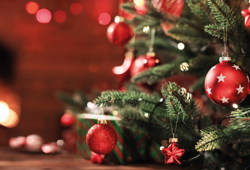 Weihnachtlich geschmückter Tannenbaum mit roten Kugeln und Sternenanhängern im rechten Bildbereich. Links im Hintergrund leuchten warme, unscharfe Lichter, die eine gemütliche Atmosphäre erzeugen.