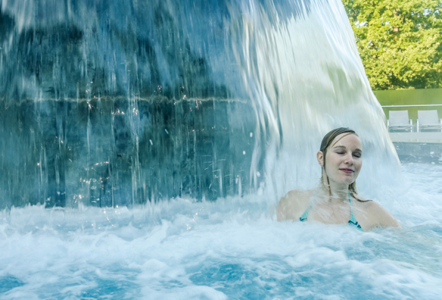 Frau entspannt im warmen Außenbecken der SaarowTherme unter einem Wasserfall, umgeben von sprudelndem Wasser und grüner Natur im Hintergrund.