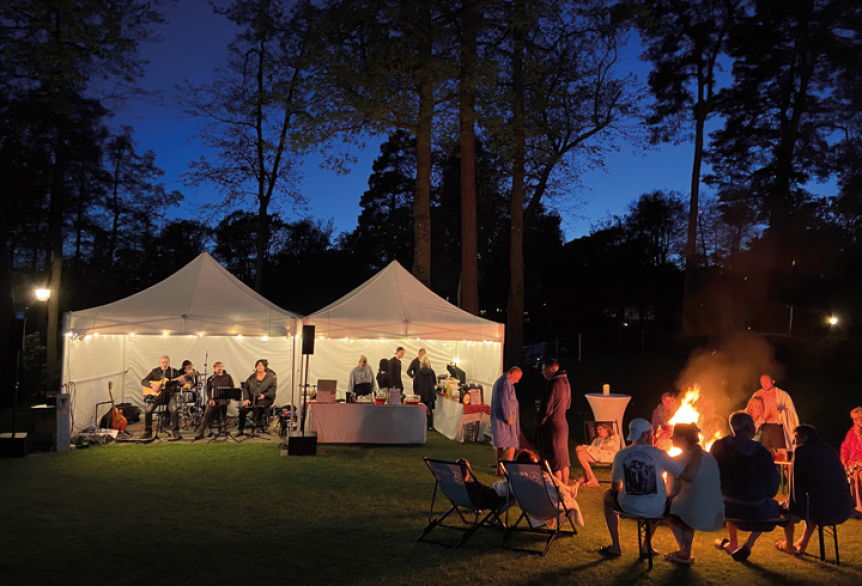 Menschen sitzen abends auf einer Wiese um ein Lagerfeuer, im Hintergrund stehen beleuchtete Zelte mit Musik.