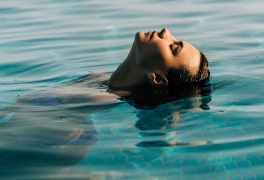 Entspannte Frau im türkisblauen Thermalsolewasser der SaarowTherme, den Kopf nach hinten geneigt, Haare im Wasser treibend, das Gesicht zur Sonne gerichtet und den Moment der Ruhe genießend.