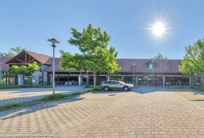 Sonniger Parkplatz vor modernem Gebäude mit Bäumen und wenigen Autos