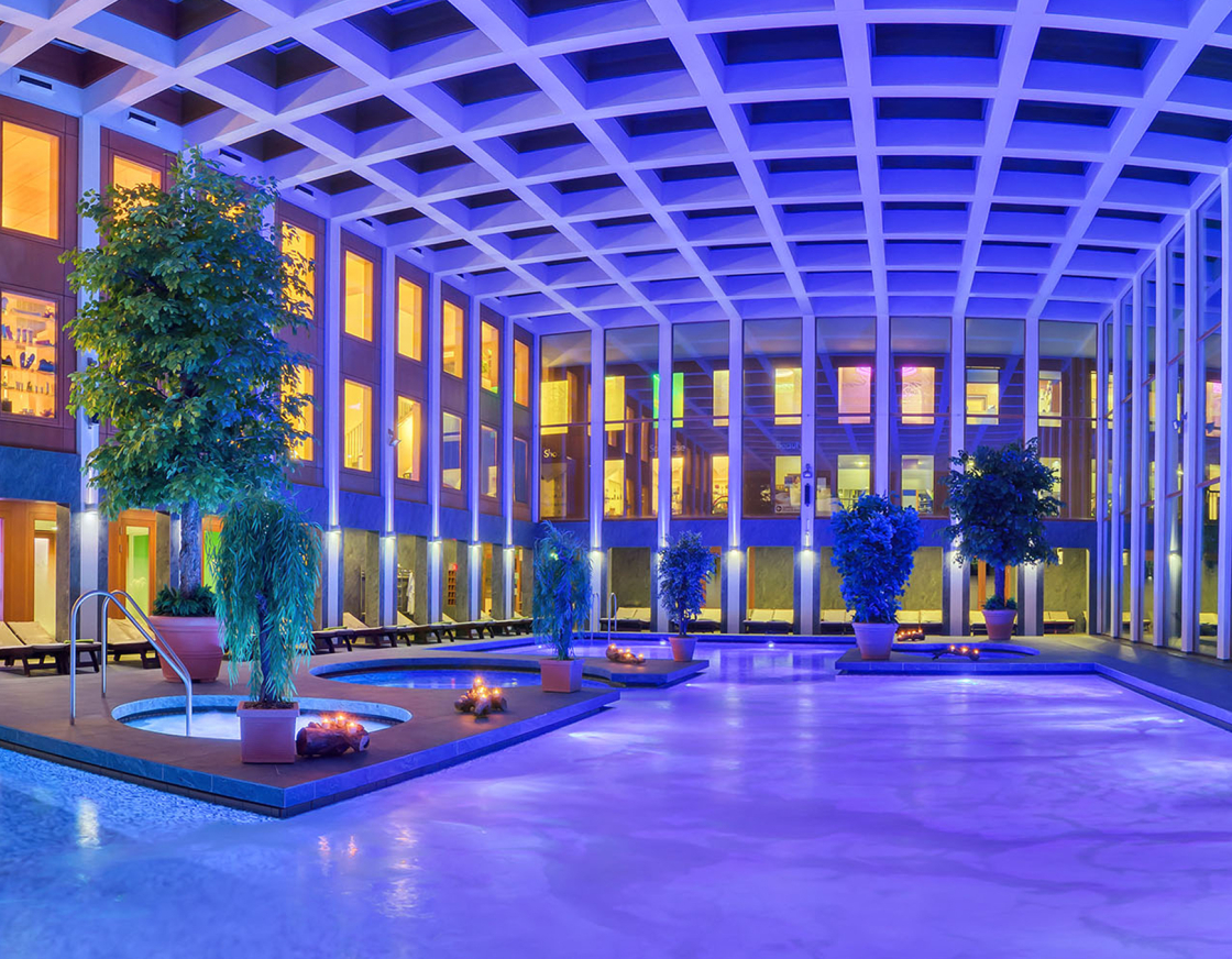 Innenbereich der SaarowTherme mit beleuchtetet Becken Abends. Dekorative Elemente wie Grünpflanzen in verschiednenen Größen und angezündete Kerzen am Beckenrand platziert. Das Solewasser ist durch die Unterwasserscheinwerfer lila gefärbt und daher wirkt das Bild blau lila. Helles bis goldenes Licht scheint durch die Glasfront vom Eingangsbereich im oberen teil des Gebäudes.