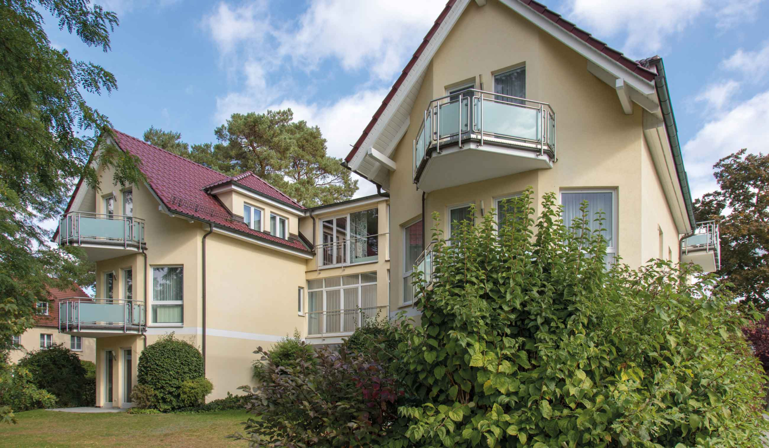 Zweistöckiges hellgelbes Wohnhaus mit roten Dächern und kleinen Balkonen, umgeben von Grünpflanzen und Bäumen unter blauem Himmel.