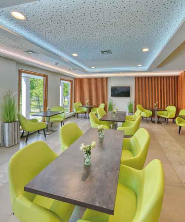 Ein großzügiger Raum mit senkrechten. mittelbraunen Holzverkleidungen als Säulen. Verteilt sind zahlreiche quadratische Tische mit dunkelgrauen Marmorplatten und grünen Sesselartigen Stühlen. Dekorative Akzente werden durch weiße Rosen in Vasen auf den Tischen gesetzt und grünen Stehpflanzen im Raum. Die Helligkeit erhält der Raum durch drei Bodentiefe Fenstern und den Deckenspots. An einer Wand hängt ein schwarzer Fernseher.