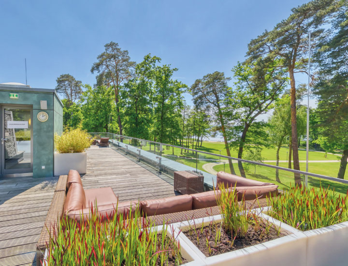 Großzügige Dachterrasse mit bequemen Lounge-Sitzgruppen und dekorativen Pflanzkästen. Die Terrasse bietet einen weiten Blick auf grüne Bäume und den nahegelegenen Scharmützelsee. Links befindet sich ein Zugang mit Glasfassade.
