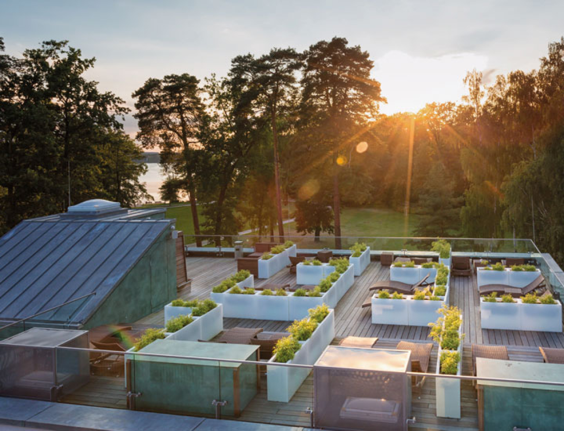 Sonnenuntergang über der begrünten Dachterrasse der SaarowTherme. In modernen Hochbeeten wachsen verschiedene Pflanzen, im Hintergrund stehen hohe Bäume, links ist ein Teil der Glasdächer des PanoramaSauna Gebäudes zu sehen.