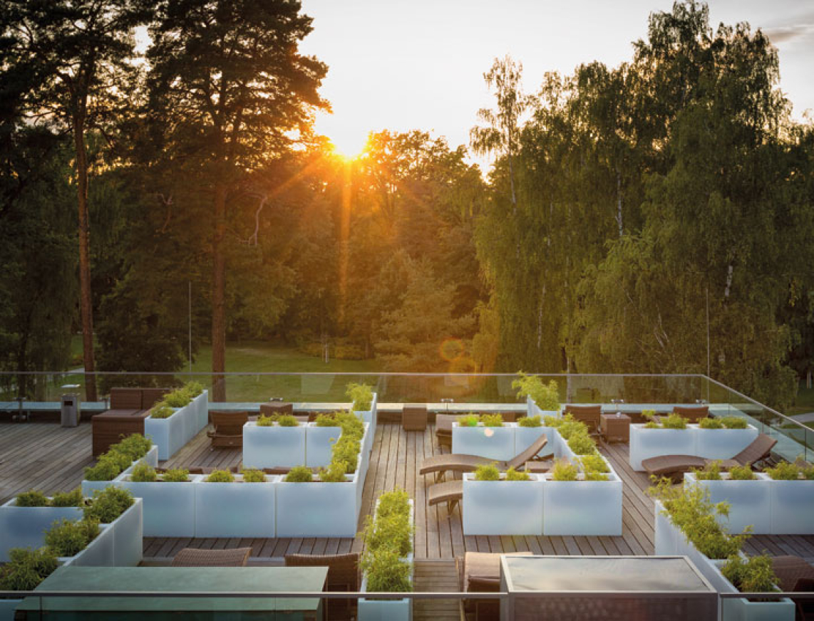 Sonnenuntergang über der begrünten Dachterrasse der SaarowTherme. In modernen Hochbeeten wachsen verschiedene Pflanzen, im Hintergrund stehen hohe Bäume, links ist ein Teil der Glasdächer des PanoramaSauna Gebäudes zu sehen.