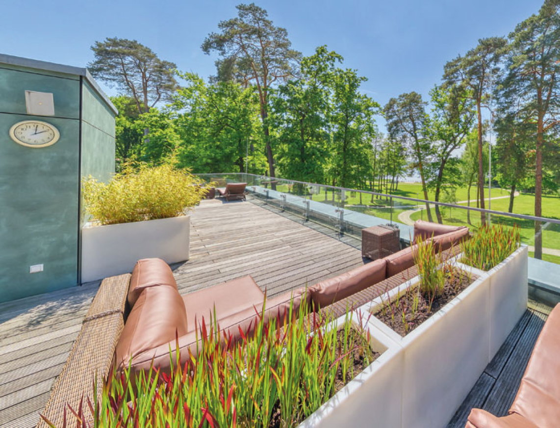 Großzügige Dachterrasse mit bequemen Lounge-Sitzgruppen und dekorativen Pflanzkästen. Die Terrasse bietet einen weiten Blick auf grüne Bäume und den nahegelegenen Scharmützelsee. Links befindet sich ein Zugang mit Glasfassade.