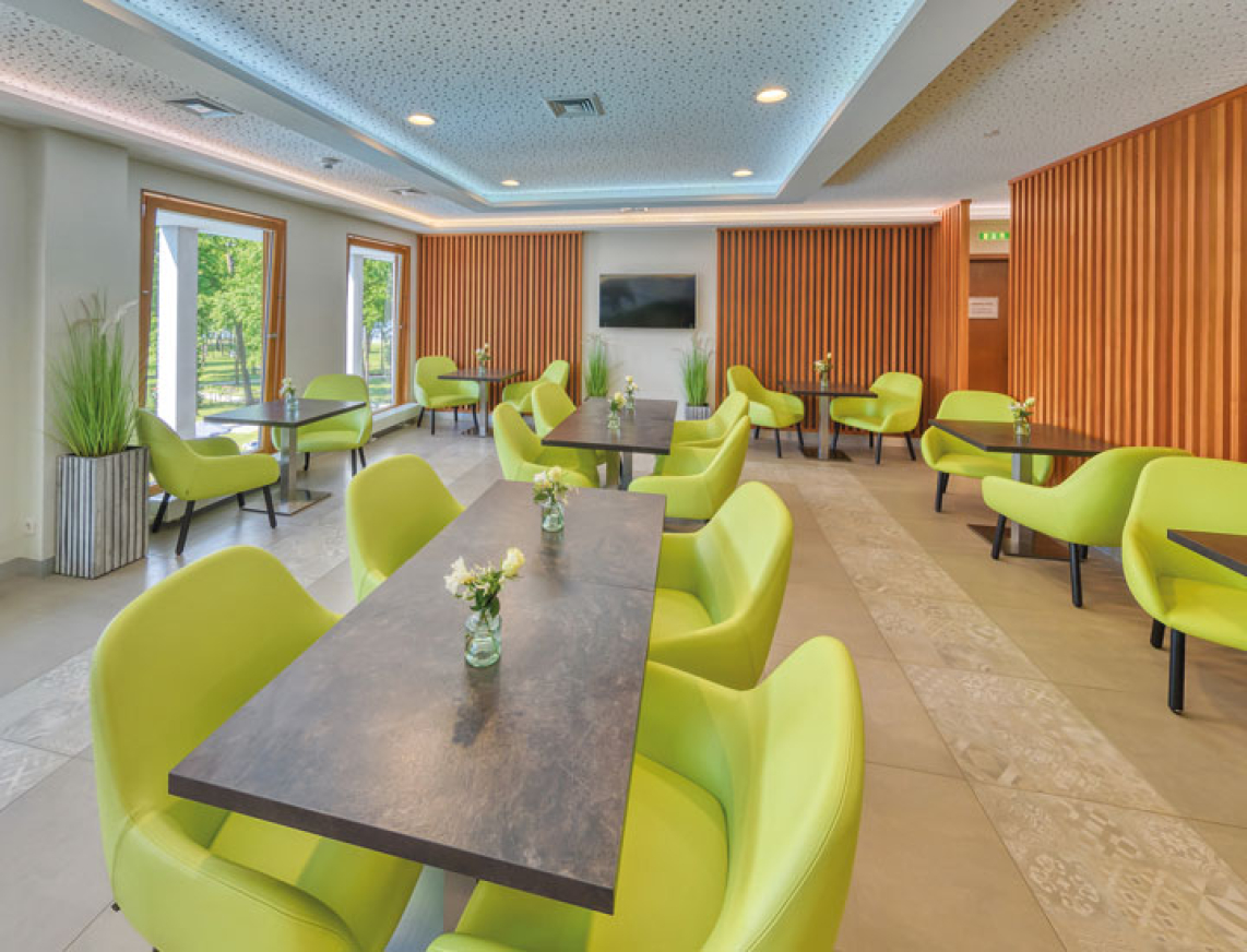 Moderner, heller Lounge-Bereich mit Limonen grünen Polsterstühlen und dunklen Tischen. Große Fenster lassen viel Tageslicht herein, während warme Holzelemente an den Wänden für eine gemütliche Atmosphäre sorgen.