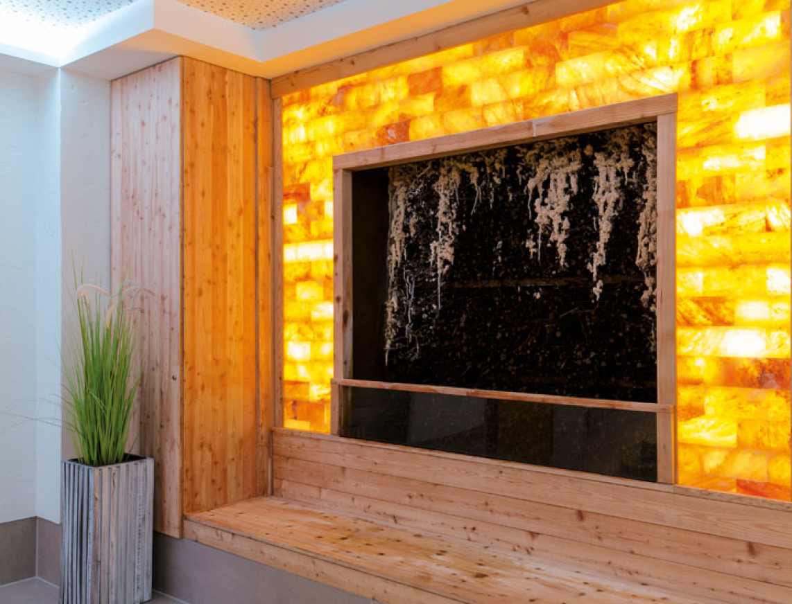 Ein Gradierwerk mit leuchtenden Salzsteinen als Umrandung. An der Sanddornreisig Wand rieselt salzhaltiges Wasser langsam herunter und erzeugt ein salziges Mikroklima zum einatmen. Vor dm Gradierwerk befindet sich eine Holzbank. Hier können sich Gäste hinsetzen und die salhaltige Luft einatmen.