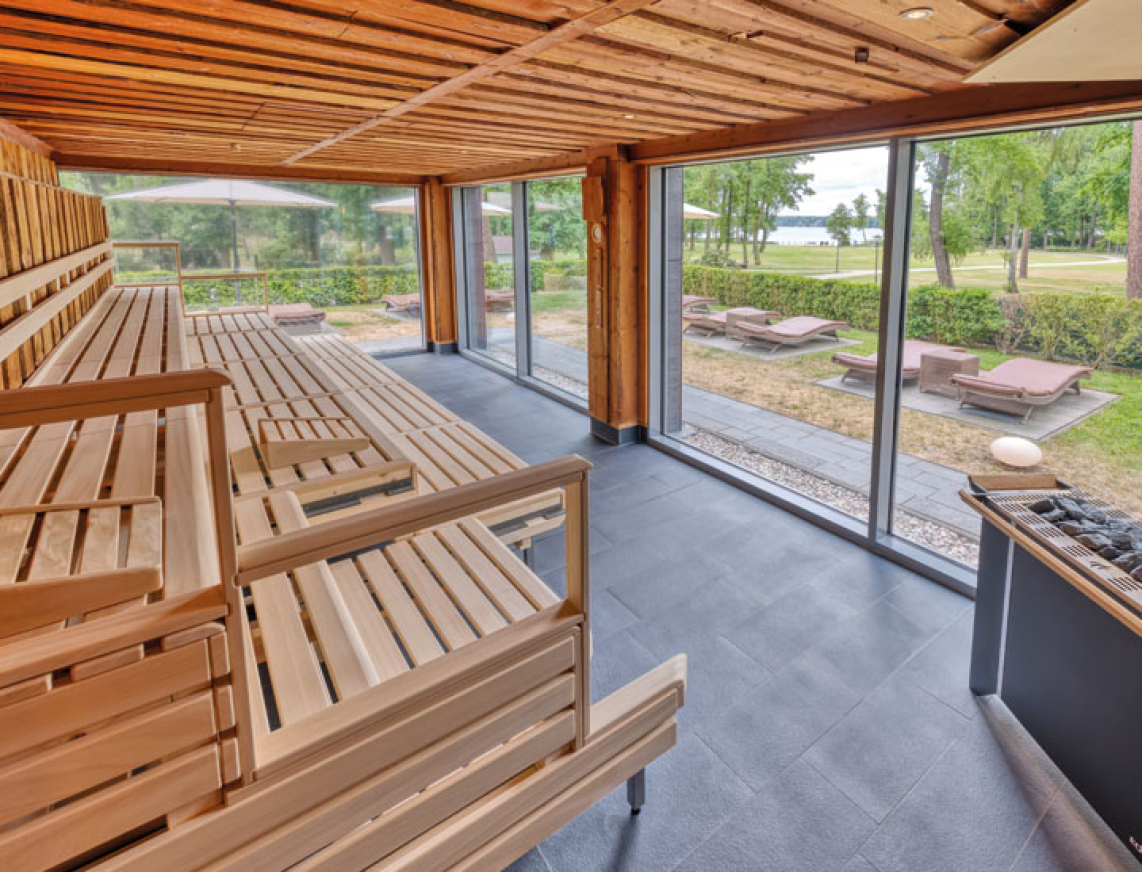 Innenansicht der großzügigen, lichtdurchfluteten KeloSauna mit Holzverkleidung und Panoramafenster. Draußen sind Liegestühle und Sonnenschirme im gepflegten Gartenbereich zu sehen.