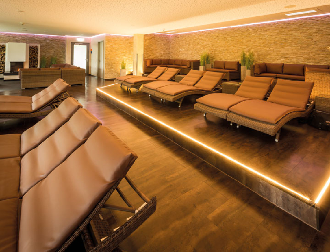 Moderner Ruhebereich mit ergonomischen, braunen Liegen auf zwei Ebenen, stimmungsvolle Beleuchtung entlang der Podeste und warme Wandgestaltung mit Natursteinoptik. Im Hintergrund Pflanzen und ein Kamin für eine entspannte Atmosphäre.