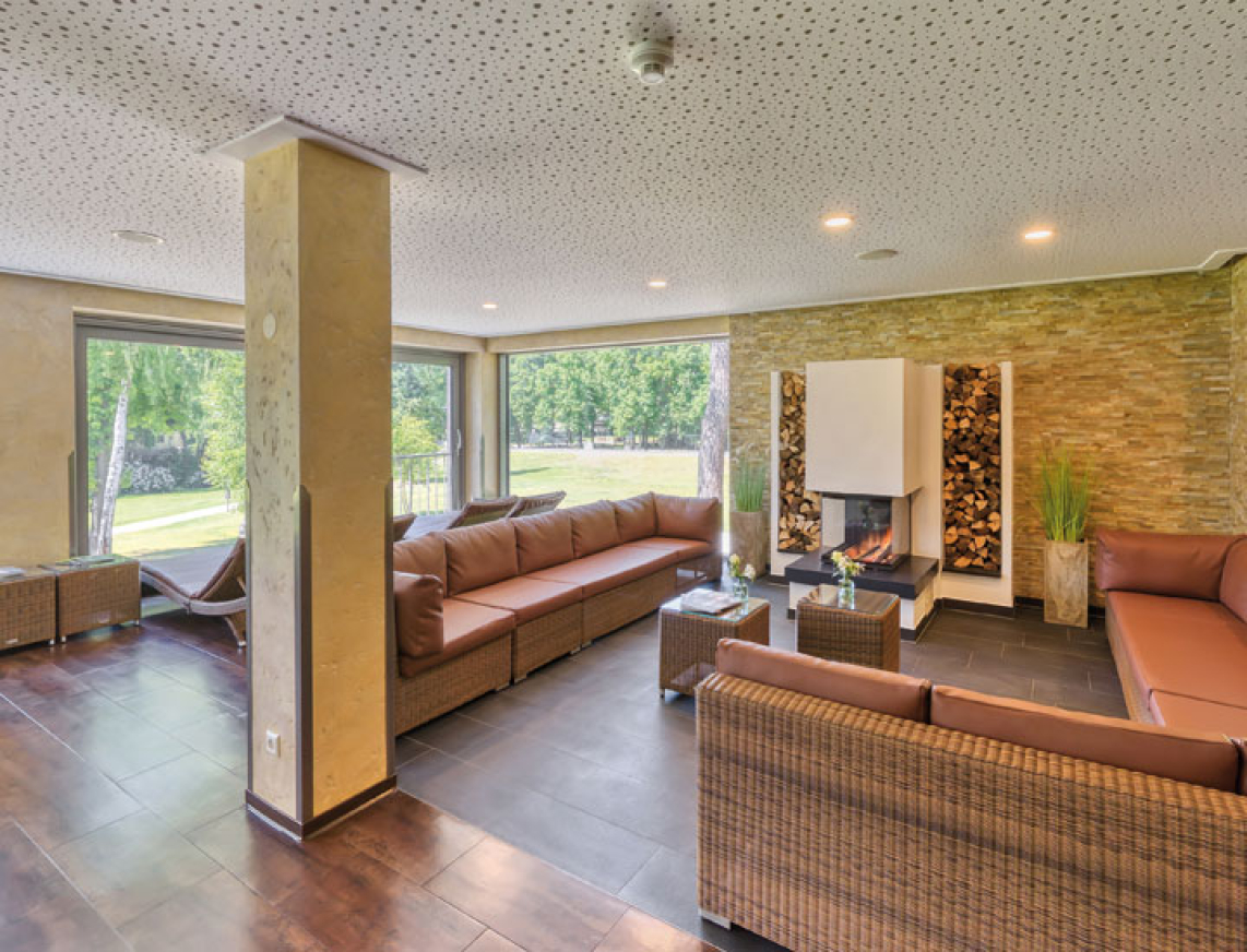 Moderner Ruhebereich mit ergonomischen, braunen Liegen auf zwei Ebenen, stimmungsvolle Beleuchtung entlang der Podeste und warme Wandgestaltung mit Natursteinoptik. Im Hintergrund Pflanzen und ein Kamin für eine entspannte Atmosphäre.