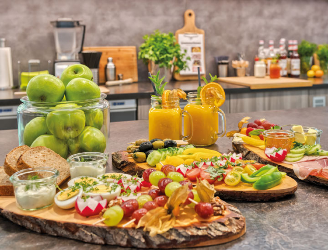 Freundlich gestaltete Saunabar mit modernem Thekenbuffet. Auf der Theke stehen bunte belegte Brote, Obst, Gemüse und frische Säfte. Dahinter sind eine gläserne Kühlvitrine mit weiteren Speisen sowie dekorative Tafeln mit Speiseangeboten an der Wand zu sehen.