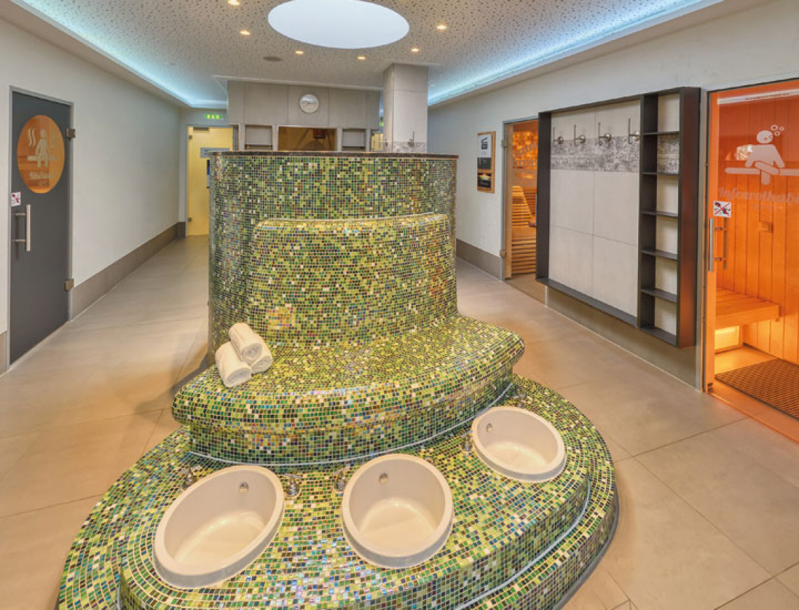 Die Saunalandschaft der SaarowTherme. Im Vordergrund sieht man drei Fußbecken mit Sitzfläche aus grünen Mosaikfliesen. Links und rechts im Bild die Eingangstüren der verschiedenen Saunen der Saunalandschaft.