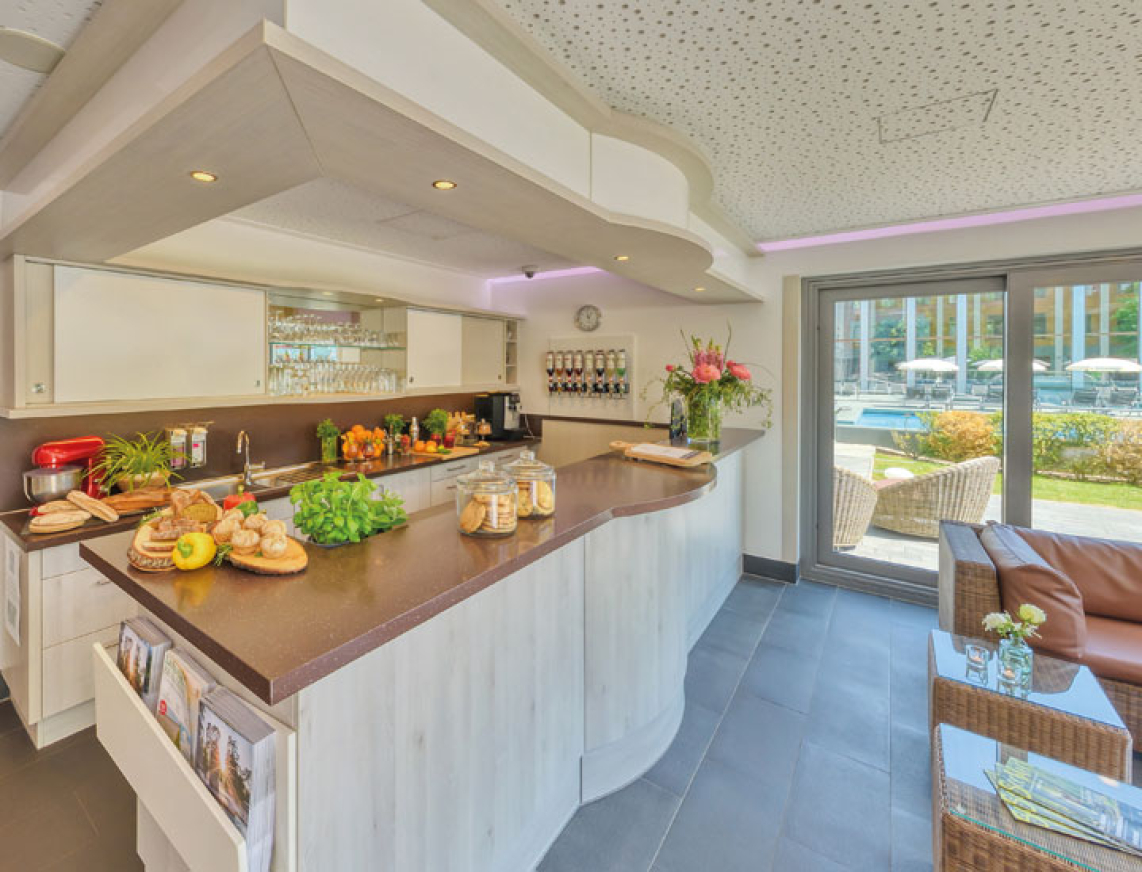 Heller Bistrobereich mit einladender Theke, auf der frische Zutaten, Obst und Getränke angerichtet sind. Links führt ein Flur in weitere Bereiche, rechts befindet sich ein gemütlicher Sitzbereich mit Blick auf die Außenanlage mit Pool.