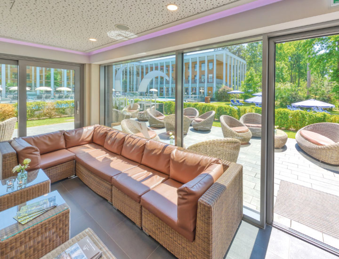 Helle Loungeecke in der PanoramaBar. Ein großes Fenster mit Blick auf die Terrasse der PanoramsSauna und dem Thermalbad im Hintergrund, sowie eine Lounge-Ecke mit bequemen Kissen schaffen eine entspannte Atmosphäre. 
