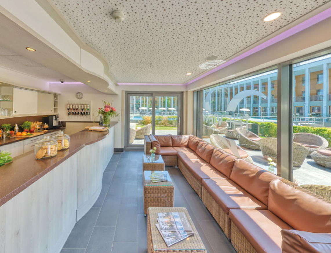 Ansicht einer Saunabar mit Loungecke am großen Fenster. An der Bartheke stehen kleine Speisen und aus dem Fenster sieht man im Hintergrund das Thermalbad der SaarowTherme.