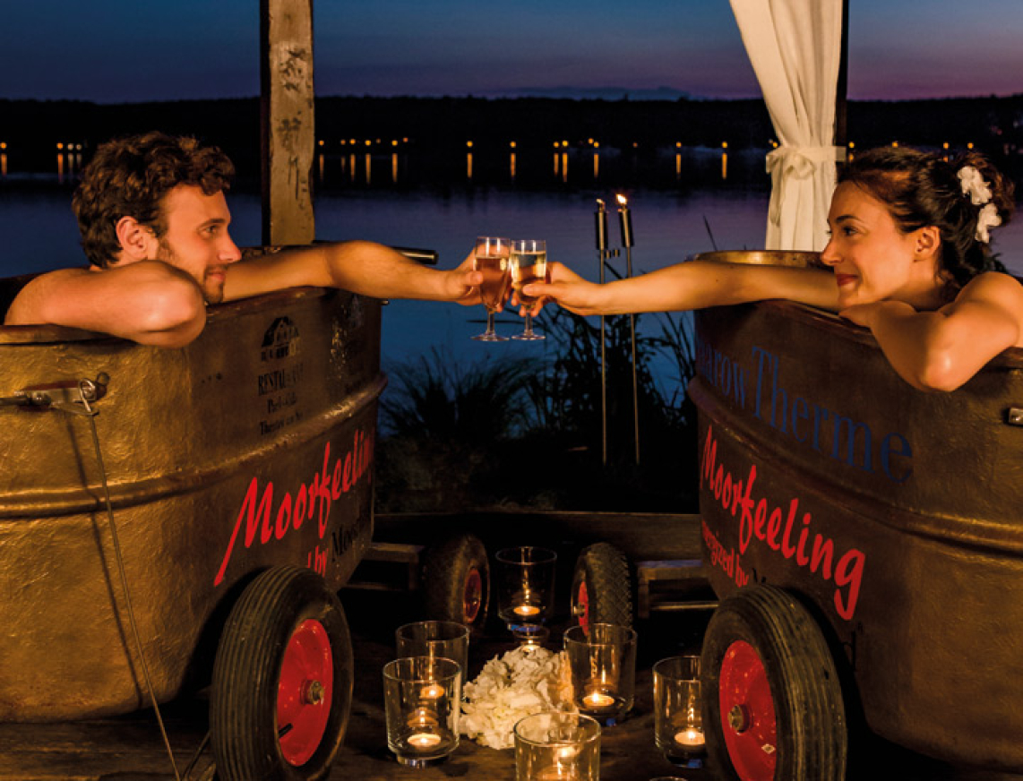 Pavillon am See mit zwei Moorwannen. Im Licht der Sterne und Kerzenschein genießt ein Pärchen ein warmes Moorbad am See. Die Wannen stehen neben einander und das Pärchen stoßend lächelnd mit Sekt zu. Zwischen den Wannen stehen auf dem Fußboden Kerzen, die für eine romantische Stimmung sorgen.