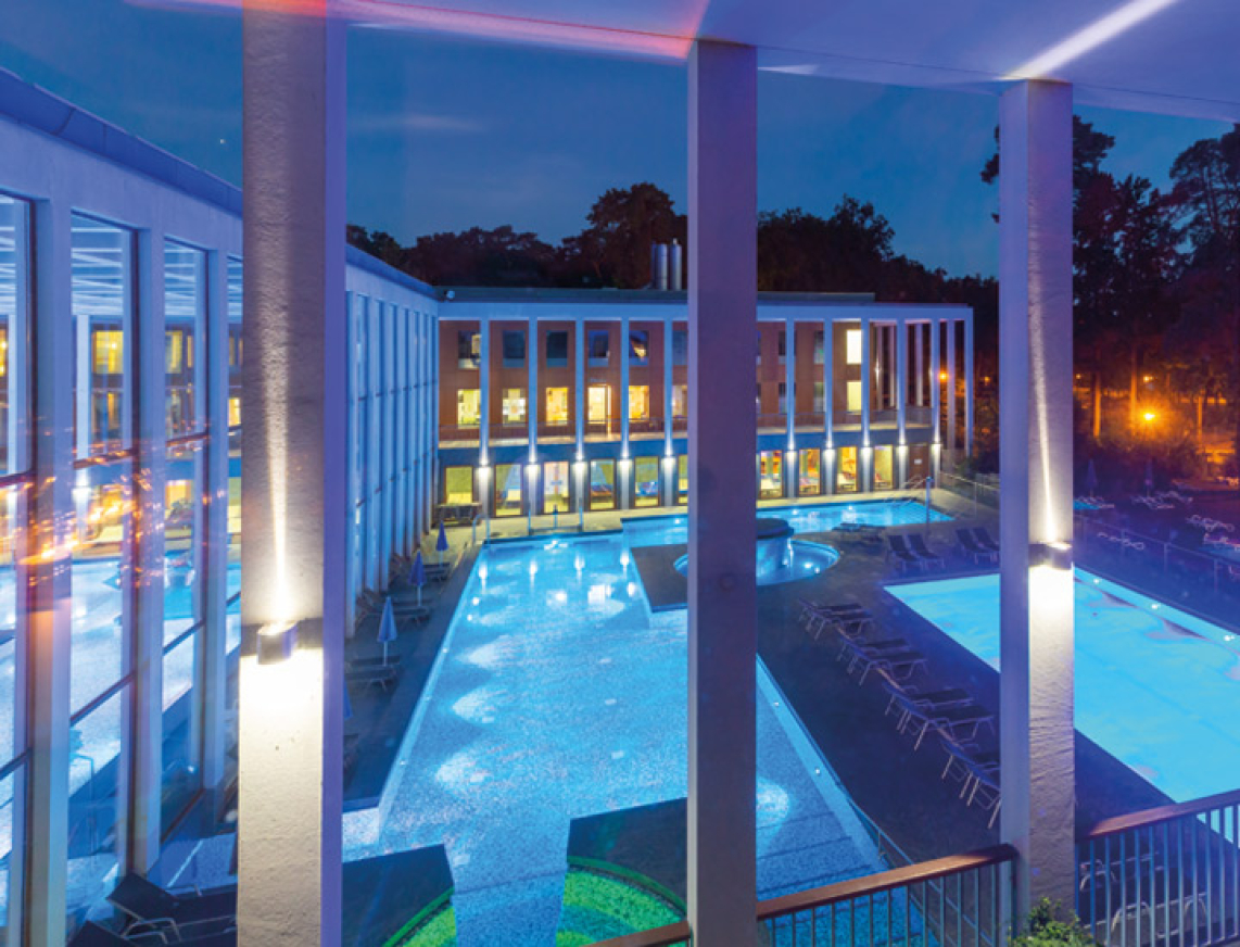 Fensteraussicht aus dem Private Spa der SaarowTherme. Aus dem Fenster erkennt man das Thermalbad der SaarowTherme mit seinen beleuchteten Becken und beleuchteten Säulen. In  blaues Licht getaucht herrscht eine romantsiche Abendstimmung.