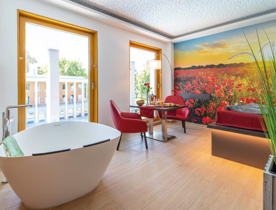 Ansicht des Private Spa der SaarowTherme. In der modern eingerichteten Suite steht links im Bild eine große Duowanne für zwei Personen. Rechts im Bild einen gedeckten Tisch mit Rosen und Sektfrühstück sowie einen in rot drapiertem Wasserbett für sinnliche Stunden.