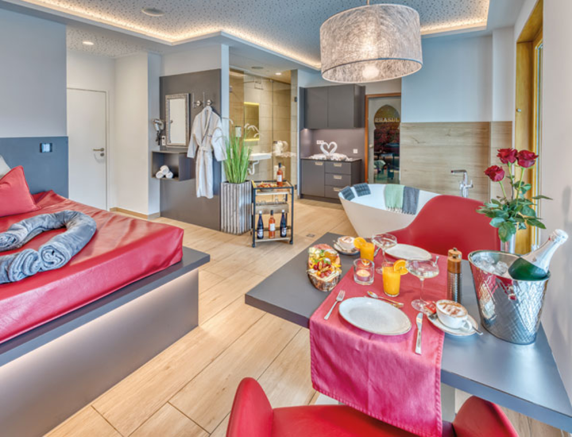 Ausschnitt aus der Private Spa Suite. Im Vordergrund steht ein mit Sektfrühstück bedeckter Tisch. Mitten im Raum steht eine freistehende Duowanne, daneben einen Rollwagen mit Getränken. Im Hintergrund erkennt man das Rhasul und den Sanitärbereich der Suite.