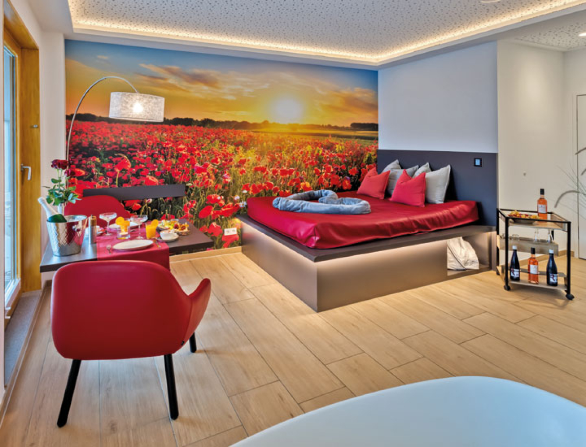 Ausschnitt aus dem Privat Spa. Ein Wasserbett mit roten Dekokissen und einer grauen Kuscheldecke. Das Bett wird von unten mit einem Lichtschlauch warm gelb beleuchtet. Auf der Hintergrundwand ist ein Sonnenuntergang zu sehen und ein Kornblumenfeld in Rot abgebildet. Zwei rote Sesselartige Stühle mit einem gedeckten Tisch.