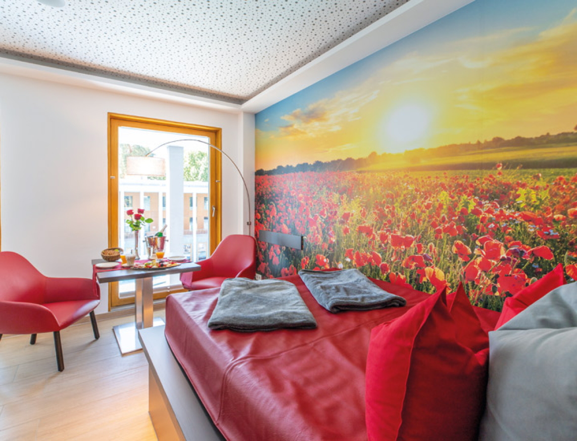 Ansicht des Private Spa der SaarowTherme. Im Vordergrund steht ein großes Wasserbet in rot drapiert mit kuscheligen Kissen und Decken. An einem großen Fenster mit Bilch auf die SaarowTherme steht ein gedeckten Tisch mit Sekt, Rosen und kleine Gerichte für eine romantische Auszeit.